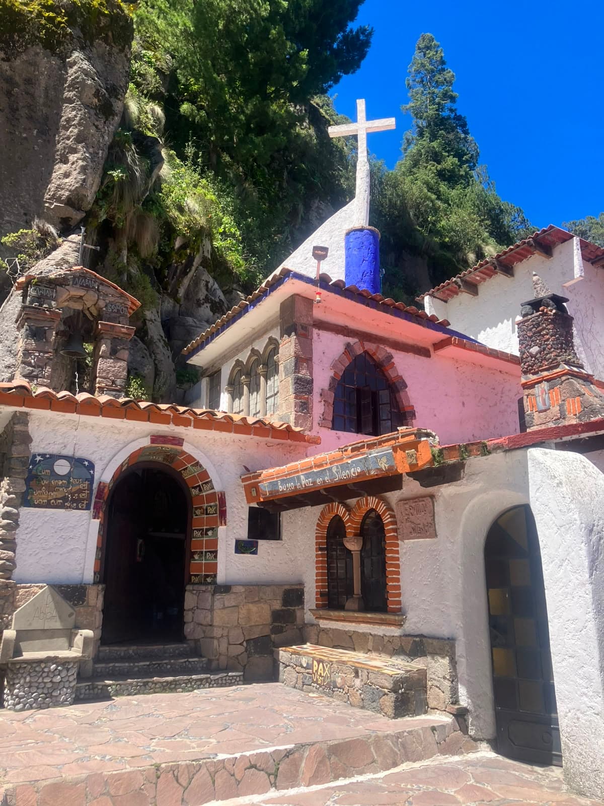 Fachada rosa y blanca de la capilla, con cruz y teja, apoyada en la roca
